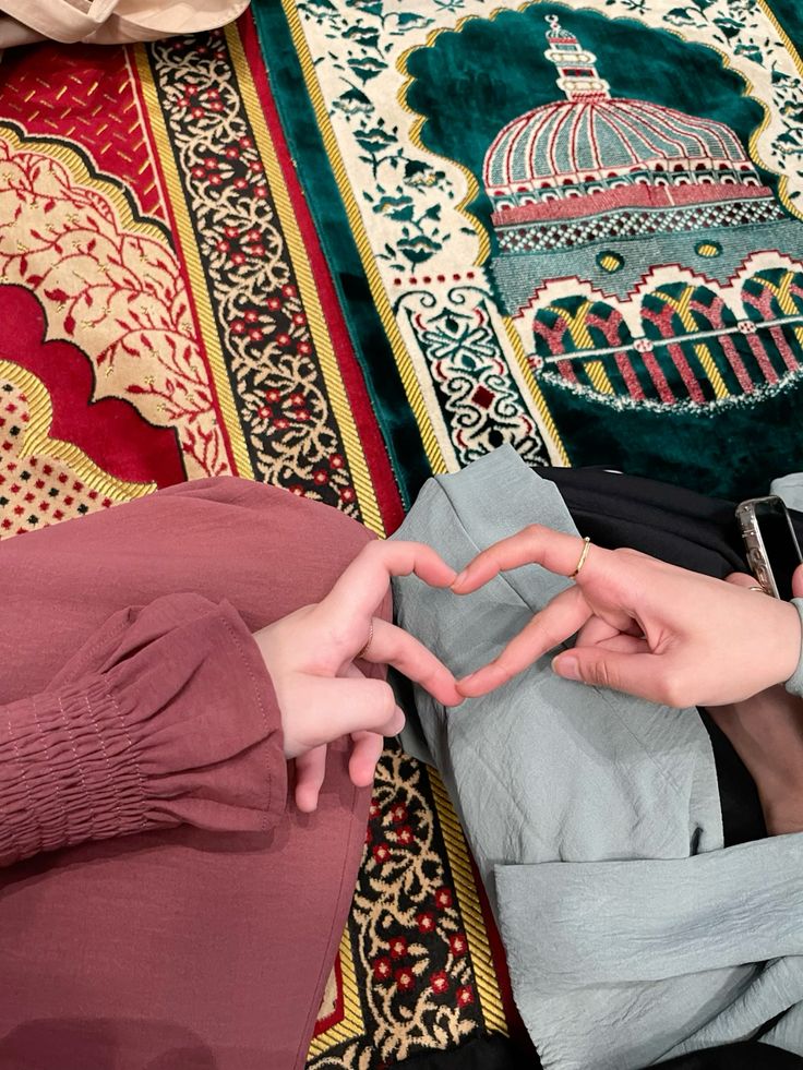 Two people sitting on a colorful prayer rug, forming a heart shape with their hands. One person wears a pink sleeve, the other a light gray sleeve, with a phone nearby. A mosque dome design is visible on the rug.
