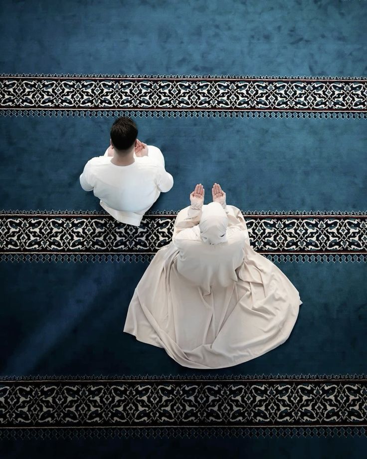 Aerial view of a man and a woman in modest clothing sitting and praying on a blue patterned carpet, likely inside a mosque.