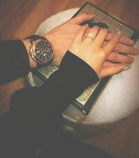 Two hands, one male and one female with wedding rings, rest on a decorative book with an ornate cover, likely a religious text. The male hand wears a watch. The lighting is soft and warm.