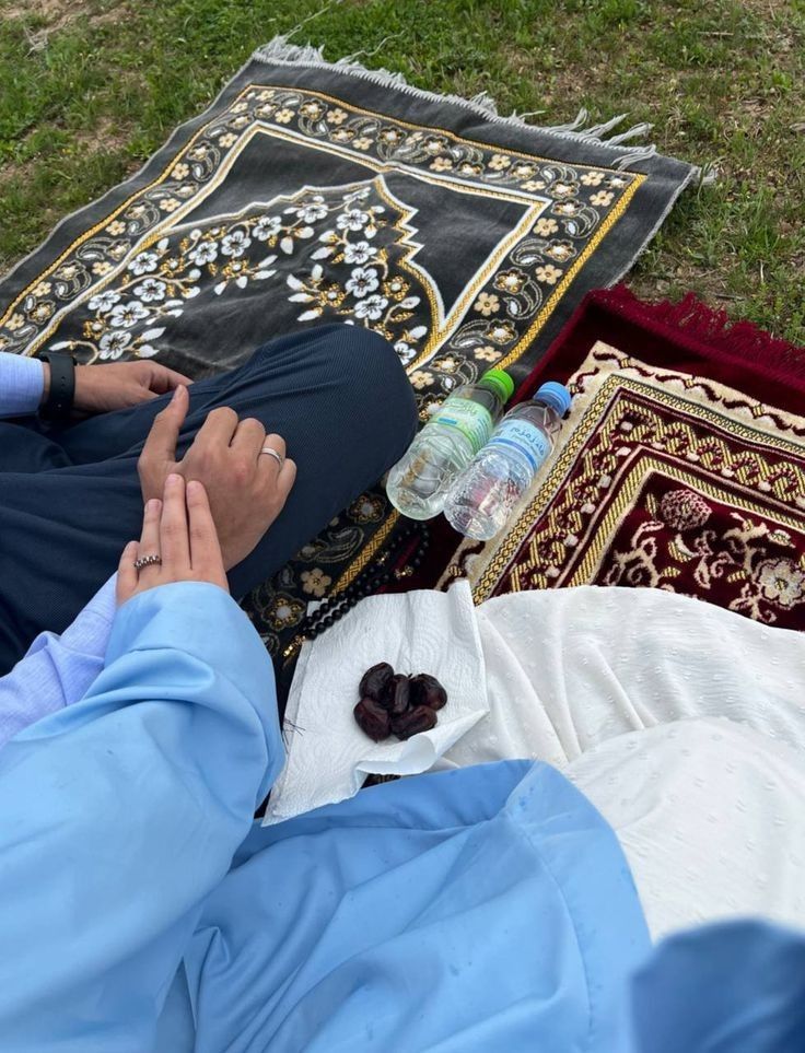 A couple holding hands while sitting on prayer mats outdoors, with water bottles and dates on a napkin beside them. The scene is set on grass, suggesting a peaceful picnic or prayer gathering.