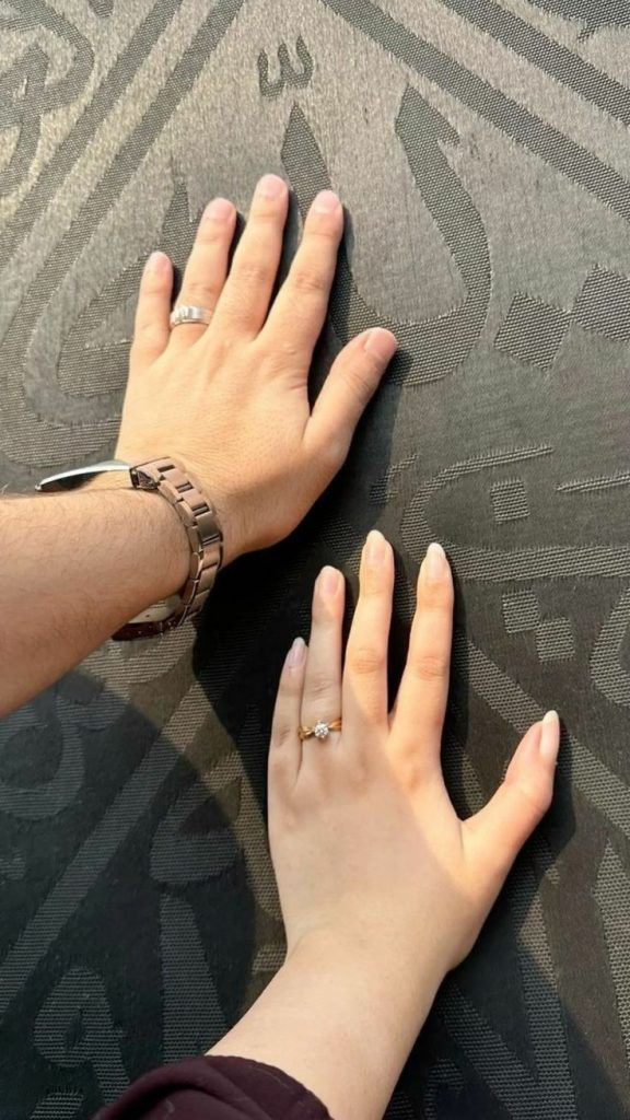 Two hands, one male with a watch and ring, and one female with a ring, touch the black cloth of the Kaaba, featuring Arabic calligraphy.
