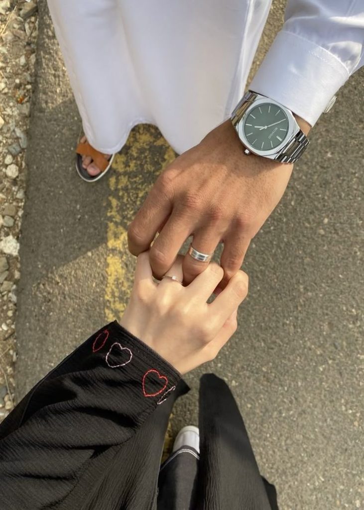 A couple holding hands outdoors, with the man wearing a watch and white traditional attire, and the woman wearing a black abaya with red heart embroidery on the sleeve. Both are wearing rings.