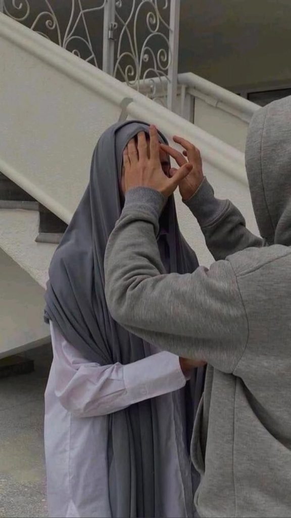 A person in a gray hoodie gently adjusts the headscarf of another person wearing a long gray hijab and white clothing, standing outside near a staircase with a decorative railing.
