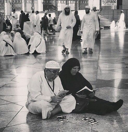 A man and woman sit on the ground reading, surrounded by others in traditional Islamic attire at a mosque. The scene is calm and reverent, with people moving and sitting in the background.