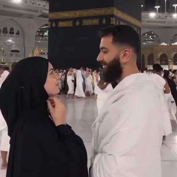 A man and woman in traditional Islamic attire stand facing each other at the Grand Mosque in Mecca, with the Kaaba visible in the background and other pilgrims nearby.