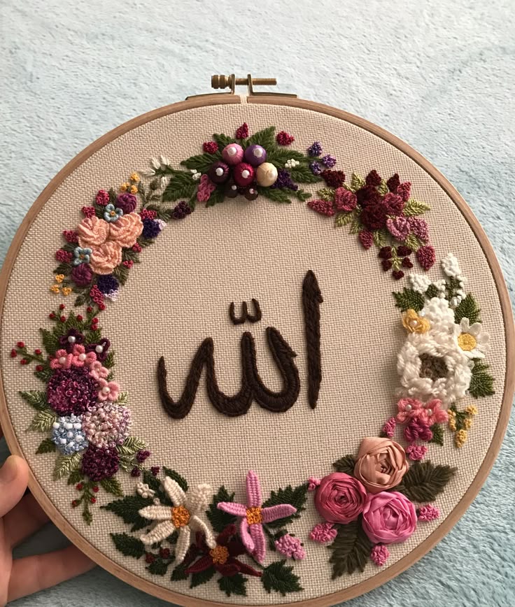 An embroidery hoop displays the word “Allah” in Arabic, surrounded by a colorful ring of hand-embroidered flowers on cream fabric. The floral design features various shades of pink, purple, red, and white.