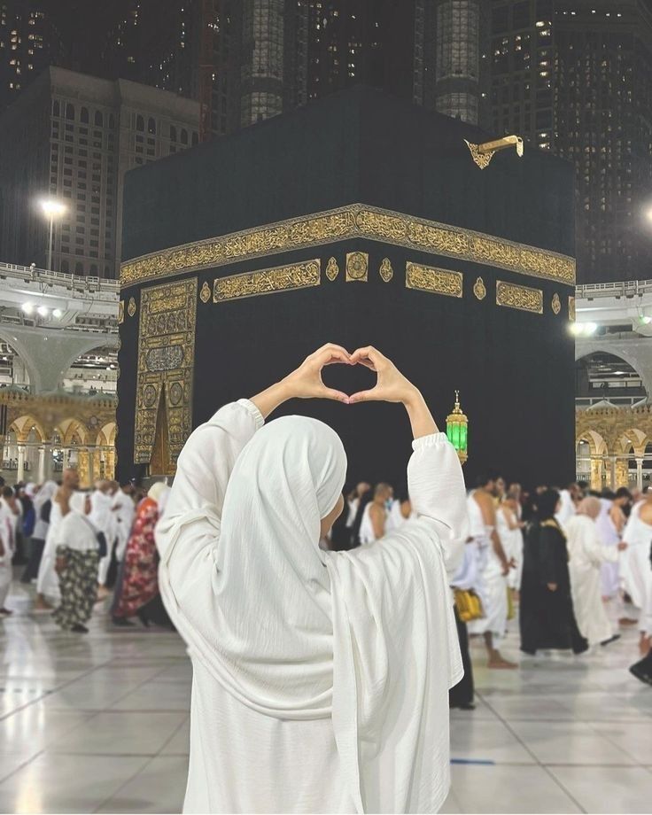 A person in white Islamic attire stands with their back to the camera, forming a heart shape with their hands toward the Kaaba in Mecca, surrounded by other pilgrims.