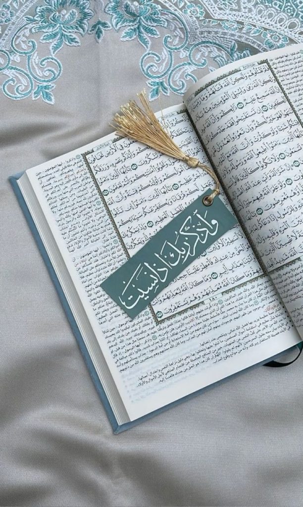 An open Quran rests on a grey, embroidered fabric. A tasseled green bookmark with Arabic script is placed on the pages. The scene evokes a sense of peace and devotion.