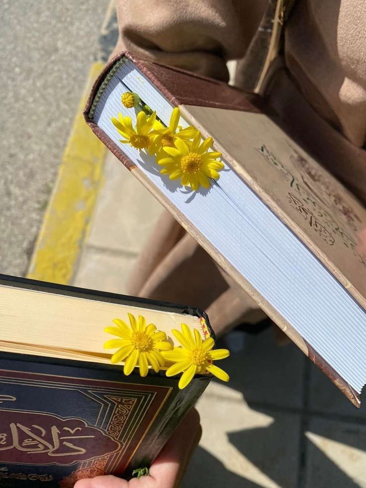 Two people hold thick books with yellow daisy-like flowers tucked between the pages as bookmarks. The books have ornate covers and are being held outdoors near a sidewalk and a yellow curb.