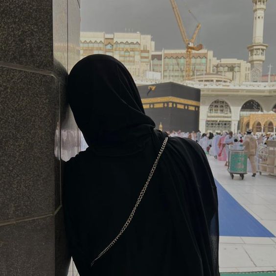 A person in a black abaya leans against a wall near the Kaaba in Mecca, with the holy site and worshippers visible in the background under a cloudy sky.