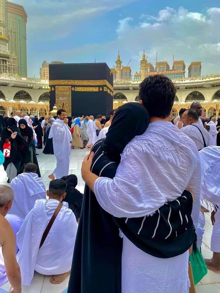 A couple embraces while facing the Kaaba at the Grand Mosque in Mecca, surrounded by other pilgrims. The sky is partly cloudy and many people are dressed in traditional white Ihram garments.